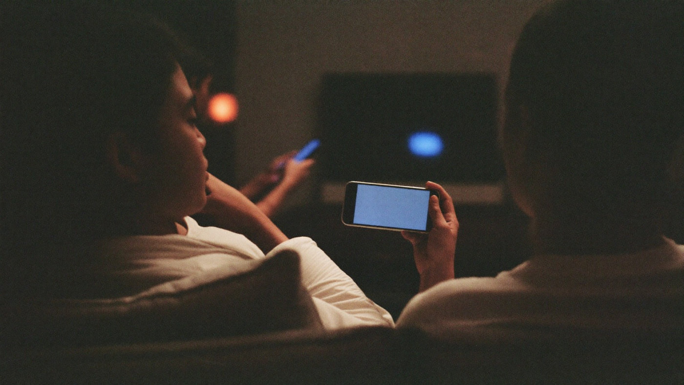 Two friends on a sofa at home watching sport with phones in their hands
